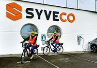 two SYVECO employees riding electric bikes in front of the building